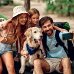 Mom and dad with their daughter with backpacks and a labrador dog take a selfie on the phone while sitting on a stone in the forest. Camping, travel, hiking.